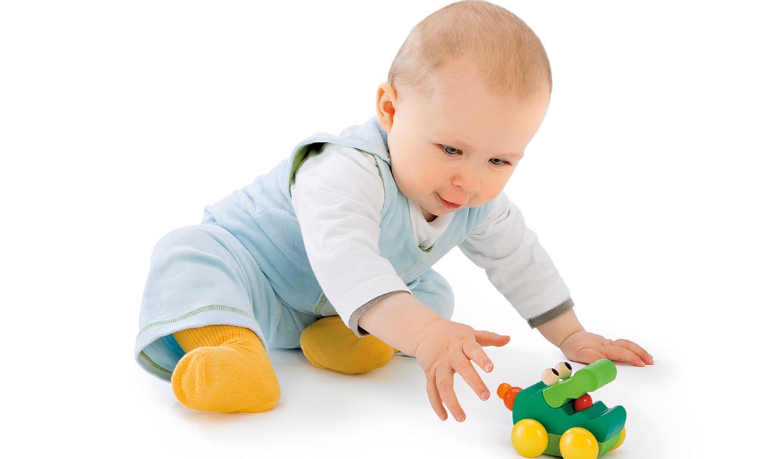 Baby im blauen Outfit sitzt auf dem Boden und greift nach einem grünen Holzspielzeugauto mit weißem Hintergrund.