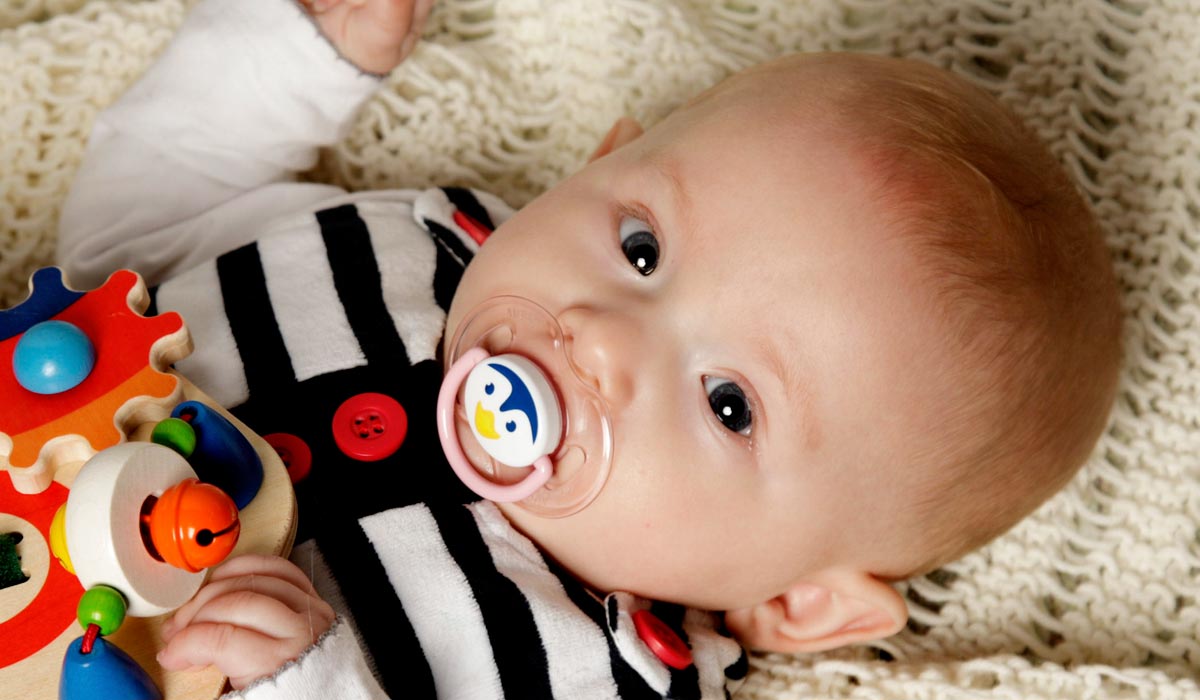 baby-sehen-betrachten Baby mit Pinguin-Schnuller auf einer Strickdecke, im gestreiften Outfit, hält ein buntes Spielzeug unter einem Spieltrapez.