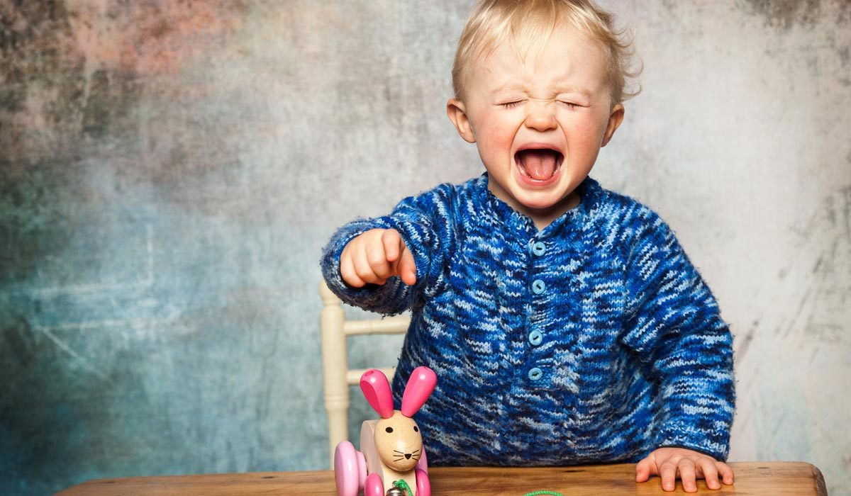 Kleinkind im blauen Pullover weint und zeigt auf ein Baby-Holzspielzeug auf dem Tisch.