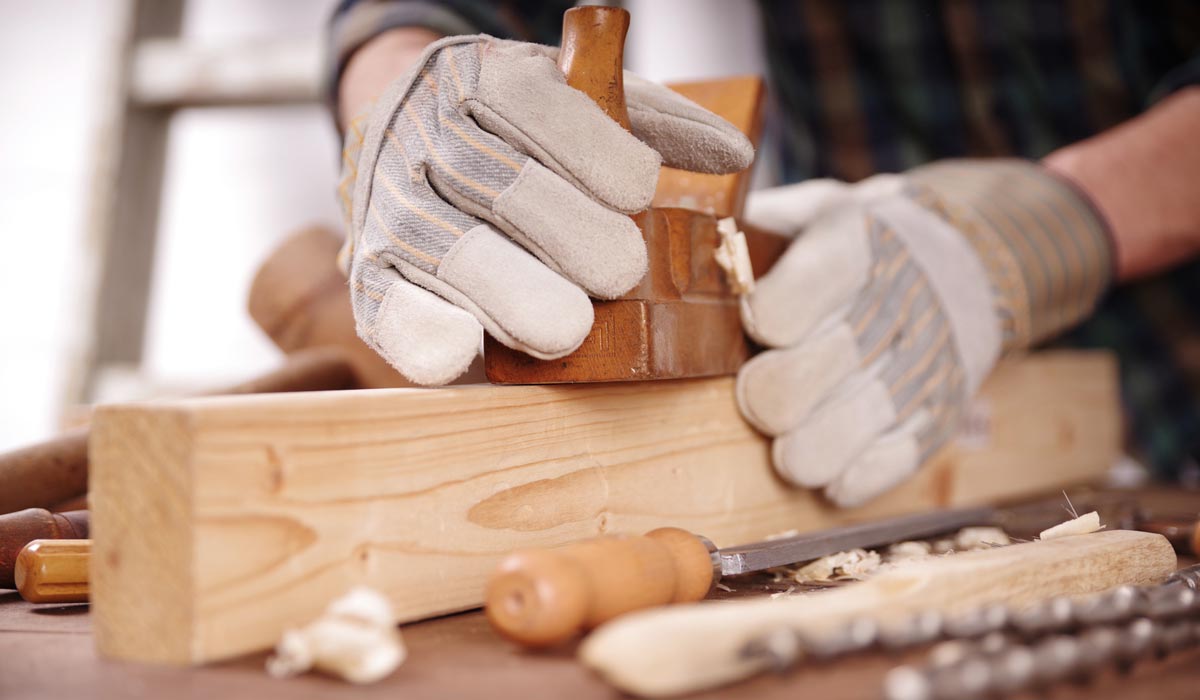 Person mit Handschuhen formt mit verschiedenen Werkzeugen in der Nähe ein Holzbrett für Baby-Holzspielzeug.