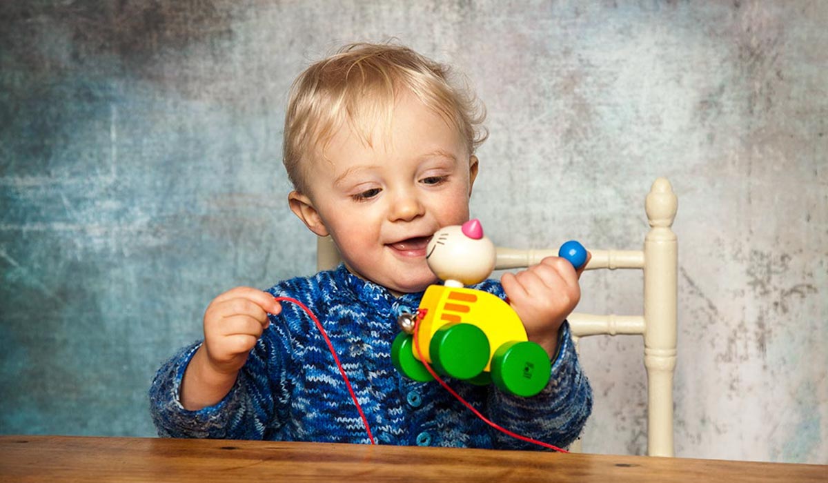 Selecta-bewegen-erleben Kleinkind im blauen Pullover spielt mit einem bunten Holzspielzeug und sitzt auf einem Holzstuhl.