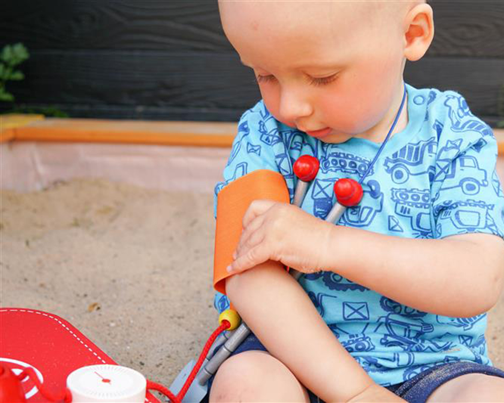 selecta-arztkoffer-holz-spielzeug-kind Ein kleines Kind überprüft seinen Arm mit einer Spielzeug-Blutdruckmanschette, die spielerisch die Sprachentwicklung fördert.