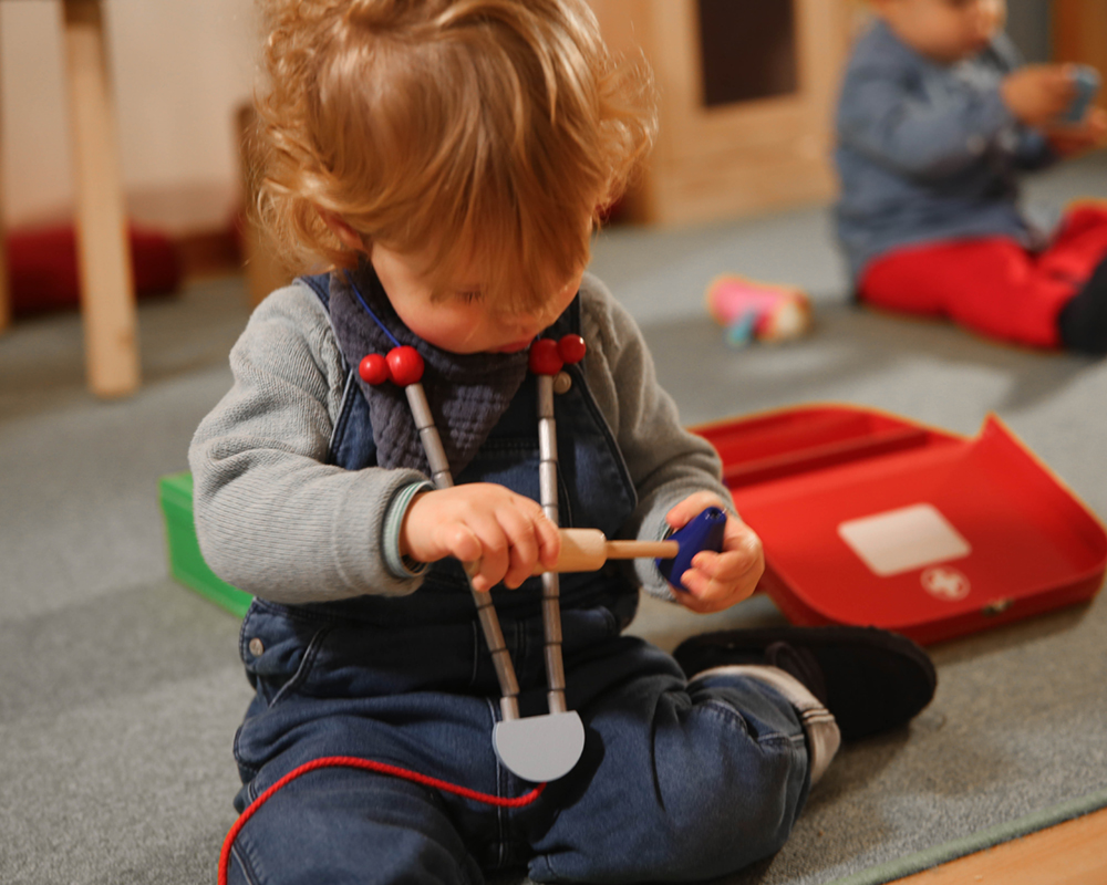 selecta-arztkoffer-holz-spielzeug-rollenspiel Kleinkind Kita: Kleinkind, das mit einem Spielzeugstethoskop spielt, ein anderes Kind ist im Hintergrund verschwommen.
