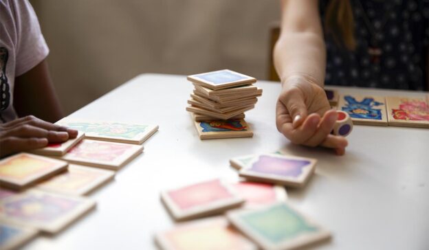 Zwei Kinder, von denen nur die Hände zu sehen sind, am Spieltisch mit Karten. Ein Kind versucht offenbar, beim Würfeln zu schummeln